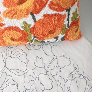 May include: A close-up of a white cushion with orange and green floral embroidery. The cushion is on top of a white canvas with a black outline of the same floral design.