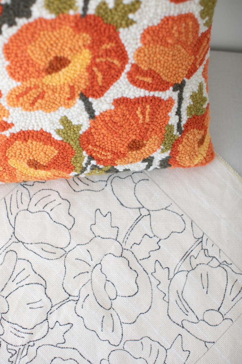May include: A close-up of a white cushion with orange and green floral embroidery. The cushion is on top of a white canvas with a black outline of the same floral design.