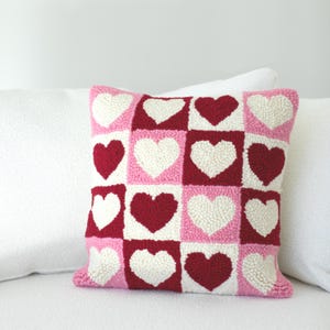 May include: A square decorative pillow with a grid pattern of hearts. The hearts are in white, pink, and red, set against a pink background. The pillow is on a white surface.