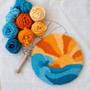 May include: A circular rug hooking project depicting a sunset and ocean scene, crafted with orange, yellow, and blue yarn. The image shows the rug in progress, alongside balls of yarn in matching colours and a rug hooking tool, all on white fabric.