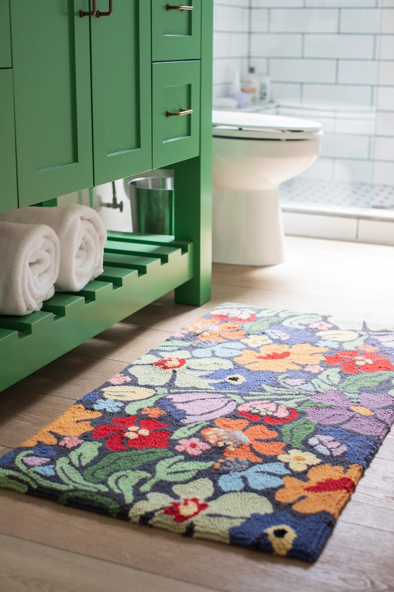 May include: A colorful floral rug with a dark blue background lies on a light wood floor. The rug features a variety of flowers in red, orange, yellow, purple, and green. A green bathroom vanity and white towels are in the background.