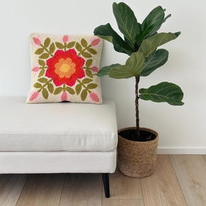 Op de afbeelding: Een decoratief vierkant kussen met een bloemenontwerp in tinten rood, oranje en geel, omgeven door groene bladeren en roze knoppen. Het kussen ligt op een lichtgekleurde bank naast een potplant met grote groene bladeren.