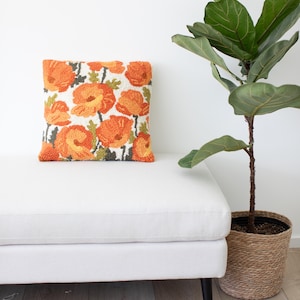 May include: A white tufted ottoman with black legs and a floral patterned cushion. The cushion features orange and green flowers on a white background. A potted plant with green leaves is in the background.