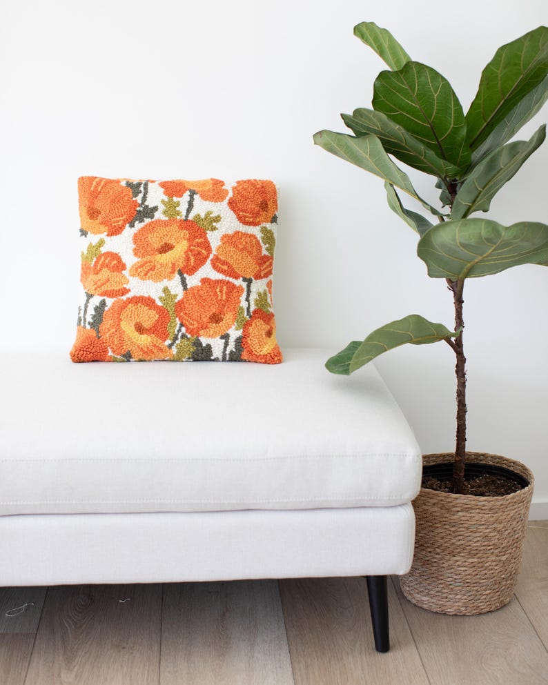 May include: A white tufted ottoman with black legs and a floral patterned cushion. The cushion features orange and green flowers on a white background. A potted plant with green leaves is in the background.