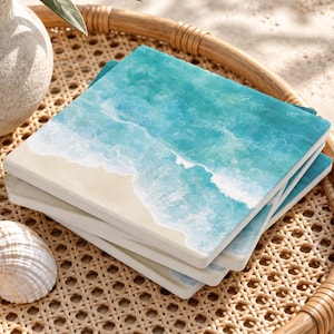 May include: A stack of square coasters featuring a printed design of a sandy beach meeting turquoise ocean waves with white sea foam. The coasters have a smooth surface and are displayed with a white ceramic vase and a seashell.