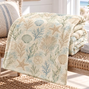 May include: A soft throw blanket featuring a coastal pattern with tan, blue, and green illustrations of starfish, seashells, sand dollars, and coral reefs printed on a cream-coloured fabric background, folded neatly over a woven chair.