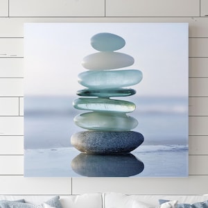 May include: A stack of smooth, light blue and gray stones balanced on top of each other. The stones are arranged in a zen-like formation, with the top stone being the smallest. The stones are set against a blurred background of a beach with blue water.