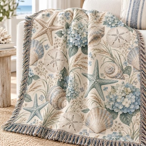 May include: A woven throw blanket featuring a coastal pattern of light blue hydrangea flowers, tan starfish, sand dollars, and various seashells on a cream background. The blanket has a decorative fringe border along the edges in blue and tan tones.