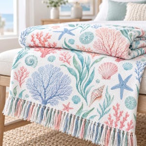 Op de afbeelding: Een witte plaid met een herhalend patroon van blauw, roze en groen zeeleven, waaronder koraal, zeesterren, schelpen en zeewier. De deken heeft een geweven textuur en is afgewerkt met lange, veelkleurige franjes aan de onderzijde.