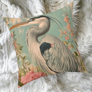 May include: Square throw pillow featuring a detailed illustration of a heron with gray and white feathers, a long orange beak, and a black and white head. The background is a light teal with pink floral accents. The pillow is on a white, textured surface.