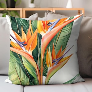 May include: A square decorative pillow featuring a vibrant watercolor illustration of a bird of paradise flower. The design showcases orange, yellow, and purple blooms with green leaves against a white background. The pillow is placed on a gray sofa.