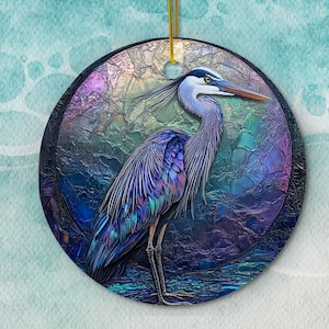 May include: A blue and purple iridescent ceramic ornament featuring a detailed illustration of a great blue heron standing on a blue and purple background.