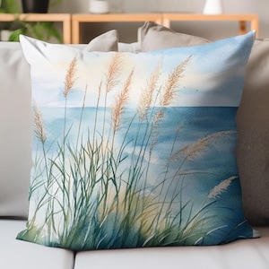 Peut inclure: Coussin carré décoratif avec une peinture à l'aquarelle d'une scène côtière. Le motif comprend de hautes herbes vaporeuses dans des tons de vert et de beige, sur fond d'océan et de ciel bleus. Le coussin est sur un canapé de couleur neutre.