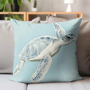 May include: A square decorative pillow featuring a detailed illustration of a sea turtle in shades of blue and white. The turtle is set against a light blue background, creating a serene and aquatic theme. The pillow is likely made of soft fabric.