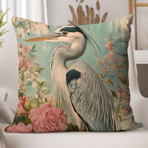 Op de afbeelding: Vierkant kussen met een gedetailleerde illustratie van een reiger. De reiger heeft een lange snavel en elegante veren, tegen een achtergrond van roze rozen en bladeren op een turquoise achtergrond. Het kussen heeft een vintage, botanisch thema.