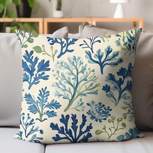 May include: Square decorative pillow with a cream background and a repeating pattern of blue and green coral and seaweed designs. The pillow is placed on a light-colored sofa.