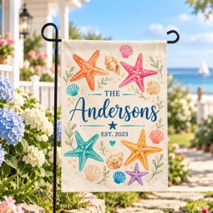 May include: A rectangular garden flag featuring a cream background with a watercolor print of colorful starfish and seashells. The text reads THE Andersons EST. 2023 in blue script and block lettering, surrounded by green leafy sprigs.