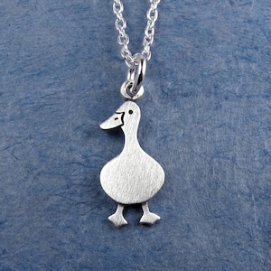 May include: A silver-toned necklace featuring a flat, stylized duck pendant with a brushed metal finish. The duck is depicted in profile, facing left, with simple etched lines for the eye and beak. It hangs from a delicate silver-toned chain.