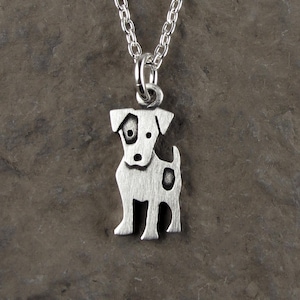 May include: A silver-toned metal pendant shaped like a small dog with a brushed finish, featuring a dark spot over one eye and another on its side. The pendant hangs from a matching silver-toned metal chain with a standard link design.