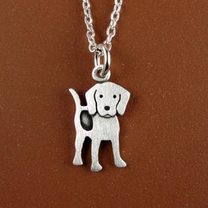 Könnte beinhalten: Eine silberfarbene Metallhalskette mit einem Anhänger in Form eines stehenden Hundes mit Schlappohren und einem dunklen Fleck an der Seite. Der Anhänger hängt an einer zarten silberfarbenen Gliederkette und hat ein matt gebürstetes Finish.