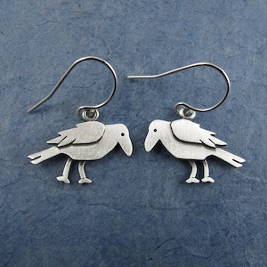 May include: A pair of silver-toned drop earrings featuring a flat, stylized bird silhouette with a matte finish. Each bird charm is suspended from a curved metal ear wire, showcasing a simple avian design with a pointed beak and two thin legs.