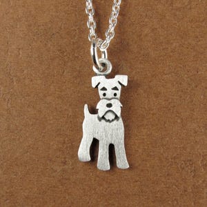 Peut inclure: Un collier en métal argenté avec un pendentif en forme de chien Schnauzer. Le pendentif présente une finition brossée avec des détails gravés pour les yeux, les sourcils et la barbe. Il est suspendu à une fine chaîne en métal argenté.