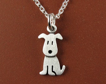 May include: A silver-toned metal pendant shaped like a stylized sitting dog with simple cut-out details for the eyes and nose. The pendant hangs from a matching silver-toned metal chain link necklace, featuring a brushed finish on the dog figure.
