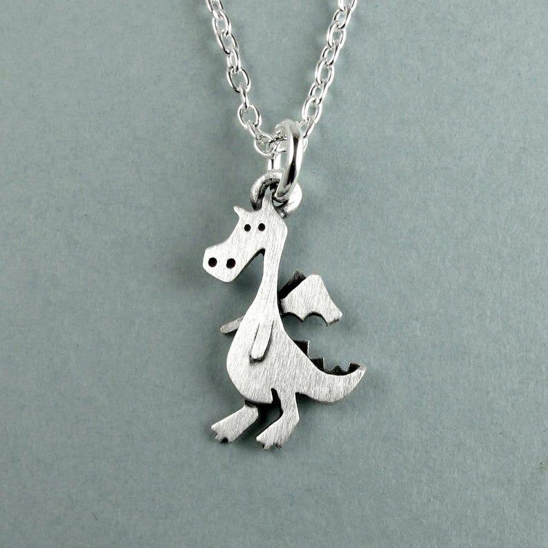 May include: A silver-toned metal necklace featuring a small, flat dragon-shaped pendant with a brushed finish. The dragon is depicted in a standing profile with a long neck, small wings, and a tail with spikes. It hangs from a delicate silver chain.