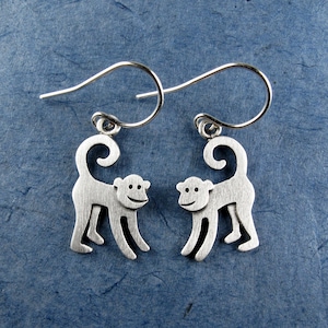 May include: A pair of silver-colored monkey earrings. Each earring features a monkey silhouette with a curved tail and smiling face. The earrings have a simple hook design for pierced ears. The background is a textured blue surface.