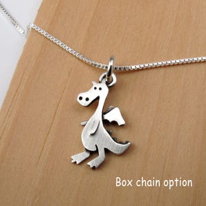 May include: A silver-toned metal dragon pendant suspended from a thin box chain necklace. The dragon charm features a flat, stylized silhouette with cut-out details for the eyes and wings. The text "Box chain option" is visible in the lower right.