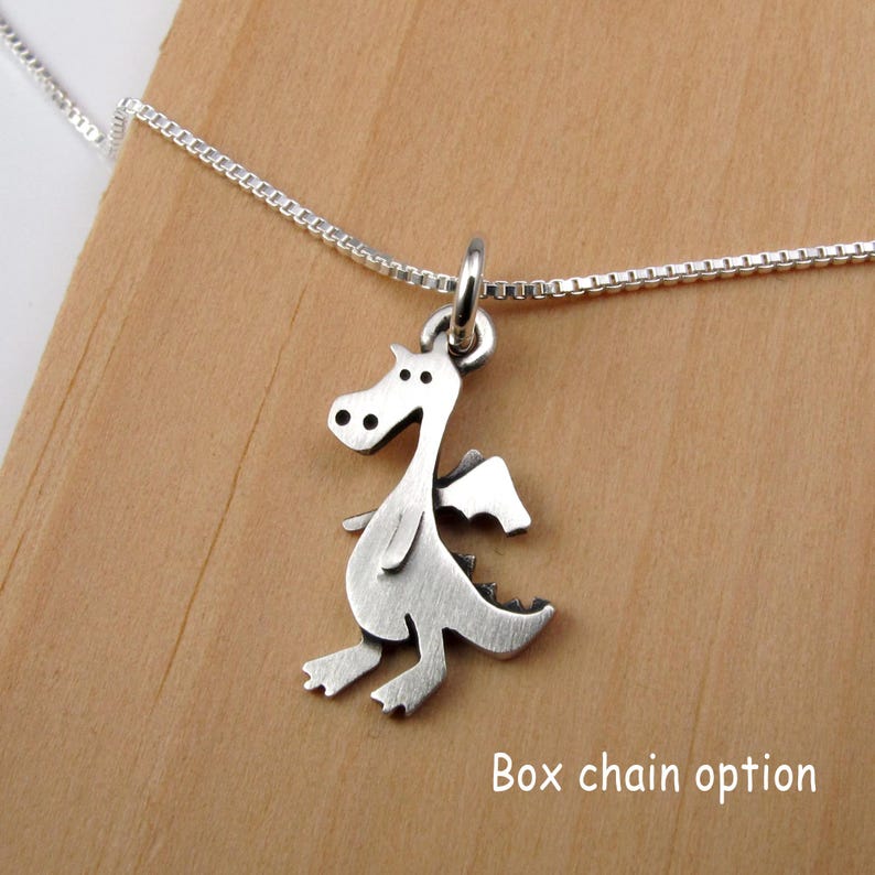May include: A silver-toned metal dragon pendant suspended from a thin box chain necklace. The dragon charm features a flat, stylized silhouette with cut-out details for the eyes and wings. The text "Box chain option" is visible in the lower right.