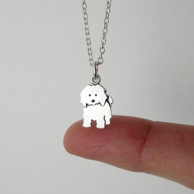May include: A silver-toned metal necklace featuring a small, flat pendant shaped like a shaggy dog. The pendant is suspended from a delicate silver-toned chain link necklace. The dog charm has simple cut-out details for eyes and a nose.