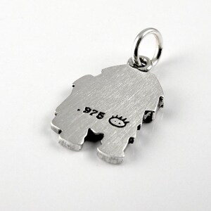 May include: A silver-toned metal charm featuring a brushed finish and a circular jump ring at the top. The surface is stamped with the text .975 and a small, stylized icon of a face with spiky hair, indicating the metal purity and maker mark.