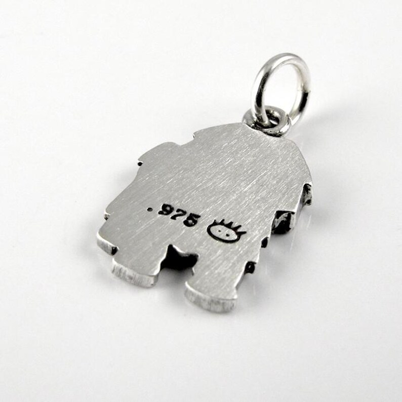 May include: A silver-toned metal charm featuring a brushed finish and a circular jump ring at the top. The surface is stamped with the text .975 and a small, stylized icon of a face with spiky hair, indicating the metal purity and maker mark.
