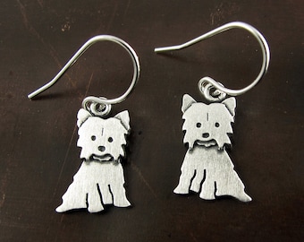 May include: A pair of silver-toned drop earrings featuring a flat, stylized silhouette of a Yorkshire Terrier dog. Each charm is suspended from a simple curved ear wire, showcasing a brushed metal finish with etched details defining the dog's face.
