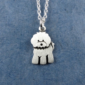 May include: A silver-coloured metal pendant shaped like a fluffy dog with a brushed finish, suspended from a matching silver-coloured metal chain. The dog charm features a rounded head, a distinct muzzle, and four legs, all rendered in a simple, flat design.