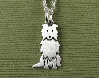 May include: A silver-toned metal pendant shaped like a sitting dog with a fluffy coat, suspended from a matching silver-toned metal chain. The pendant features a brushed finish and simple cut-out details for the eyes and nose on the dog's face.