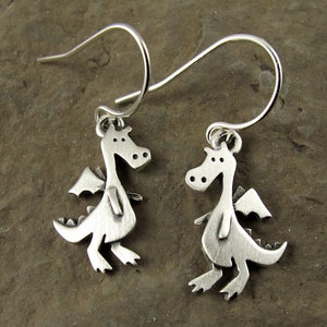 May include: A pair of silver-colored dragon earrings. Each earring features a small, stylized dragon with wings, legs, and a tail. The dragons are suspended from curved ear wires.
