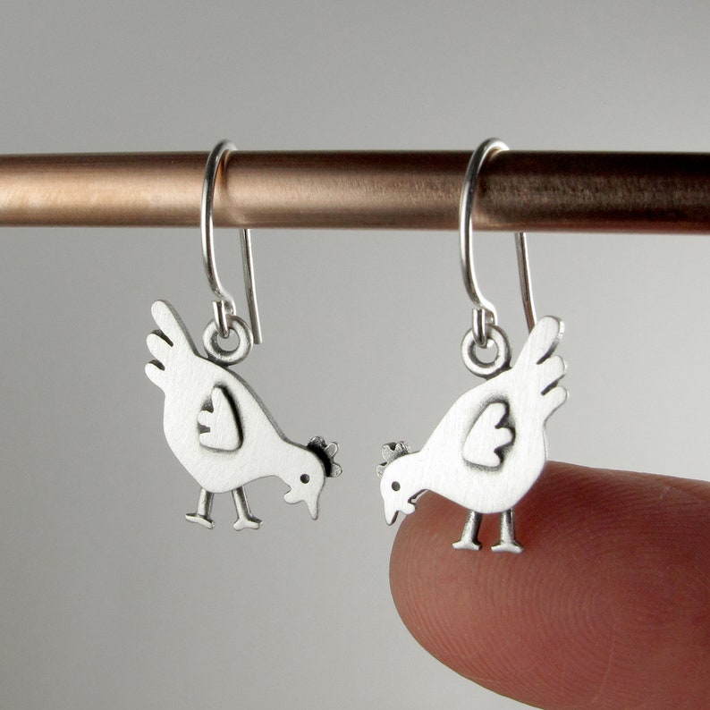 May include: A pair of silver-toned drop earrings featuring a flat, stylized chicken silhouette with a matte finish. Each earring hangs from a simple curved ear wire, depicting a bird with a small wing, a comb, and two thin legs.