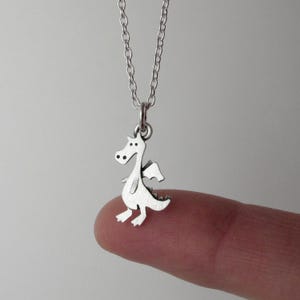 May include: A silver-toned necklace featuring a small, flat, cartoon-style dragon pendant. The dragon is depicted in profile with simple cut-out details for the eyes, wings, and tail. The pendant hangs from a delicate, thin silver-toned metal chain.