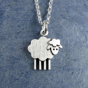 May include: A silver pendant necklace with a sheep charm. The sheep has a fluffy body and a detailed head with two small dots for eyes. The pendant hangs from a delicate silver chain. The background is a textured blue.