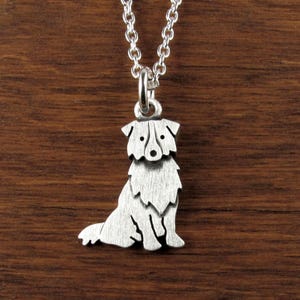 May include: A silver-colored pendant necklace featuring a detailed silhouette of a dog. The dog is sitting, with its head and body clearly defined. The pendant hangs from a delicate silver chain, creating a simple yet elegant accessory.