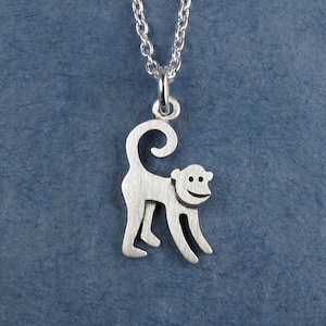 May include: A silver-colored pendant necklace featuring a stylized monkey design. The monkey has a curved tail and a smiling face. The pendant hangs from a delicate silver chain, creating a playful and charming accessory.