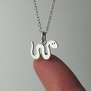 May include: A silver-coloured necklace featuring a small, flat, snake-shaped pendant with a smooth, matte finish. The snake has a curved, undulating body and two small, dark, circular indentations representing eyes. It hangs from a delicate chain.