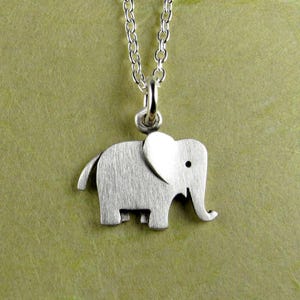 May include: A silver-colored elephant pendant hangs from a delicate chain. The pendant features a stylized elephant with a textured surface and a small, round eye. The chain and pendant are made of a shiny, metallic material, creating a simple, elegant necklace.