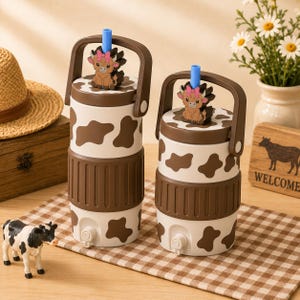 May include: Two cylindrical beverage dispensers with a white and brown cow print pattern. Each features a brown carrying handle, a blue straw, and a small brown figurine of a highland cow with a pink bow on top. A small spigot is at the base.