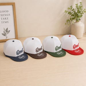 Peut inclure: Quatre casquettes de baseball blanches avec des visières colorées bleu marine, marron, vert et rouge. Chaque casquette présente le mot Dad en écriture cursive de couleur assortie et un emblème circulaire Best Dad Ever sur la visière.
