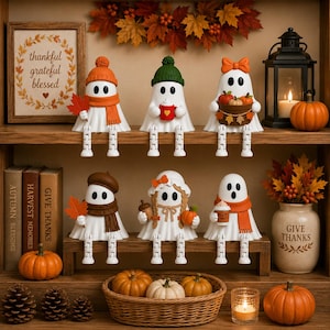 May include: Six white ghost figurines with dangling legs arranged on shelves. The ghosts wear autumn-themed accessories like hats, scarves, and hold items including maple leaves, mugs, and pumpkins. The display includes a sign reading thankful grateful blessed, a lantern, books, pinecones, and pumpkins.