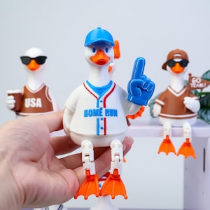 May include: A white rubber duck figurine wearing a blue baseball cap and a white baseball jersey with blue and red trim. The jersey features the text HOME RUN in blue. The duck holds a blue foam finger and has articulated white legs with orange feet.