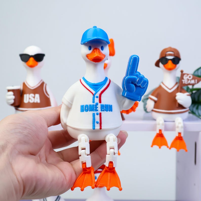 May include: A white rubber duck figurine wearing a blue baseball cap and a white baseball jersey with blue and red trim. The jersey features the text HOME RUN in blue. The duck holds a blue foam finger and has articulated white legs with orange feet.
